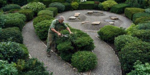 Person pruning shrubs in an offset garden layout where deep beds alternate left and right across a gravel path